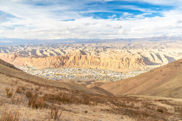 A stunning landscape featuring a small urban settlement nestled between vast, golden desert ridges under a vivid blue sky with scattered clouds. The hills contrast with the intricate natural formation