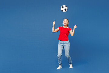 Full body young happy cheerful fun cool smiling woman fan wear red t-shirt cheer up support football sport team hold in hand toss up soccer ball watch tv live stream isolated on plain blue background.