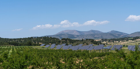 a vast solar farm with rows of solar panels spread across the landscape. The panels are surrounded by green fields and distant mountains under a clear blue sky.