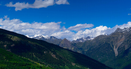 A stunning landscape showcasing a range of majestic mountains with vibrant green forests under a clear blue sky, dotted with fluffy white clouds.