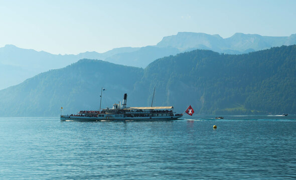 A vintage paddle steamer boat sails across a calm lake with Swiss mountains in the background, featuring a Swiss flag and a buoy toward a serene landscape.