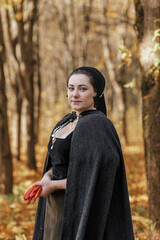 Young woman in medieval dress and cloak standing by tree in forest. Middle Ages