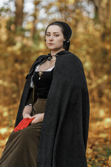 Young woman in medieval dress and cloak standing by tree in forest. Middle Ages