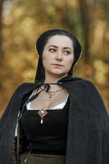 Young woman in medieval dress and cloak standing by tree in forest. Middle Ages