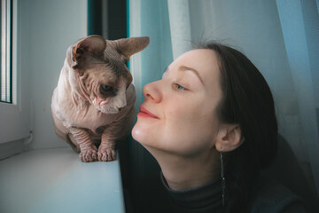 Cat and young woman affection. Portrait of a brunette girl with Canadian sphinx cat indoors. A...