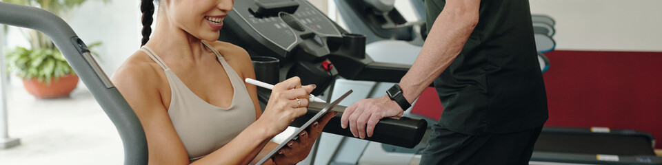 Smiling trainer holding a tablet, recording client workout data in gym. Client standing beside, wearing fitness attire, while receiving feedback on exercise routine