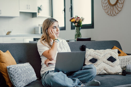 One caucasian unhappy girl looking outside sitting on sofa using laptop computer thinking on problems. Female worried woman studying and working.