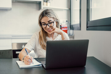 Millennial blonde girl sit at desk in living room study on laptop making notes, concentrated young woman work on computer write in notebook, take online course or training at home, education concept.