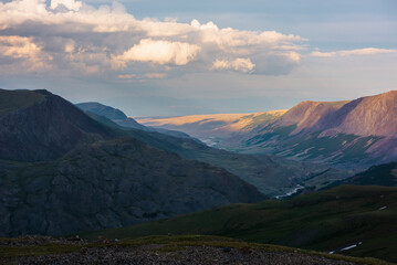 Dramatic aerial top view to golden steppe illuminated by setting sun and mountain range silhouette under clouds in gold sunset tones. Evening alpine landscape under cloudy sky in vivid sunset color.