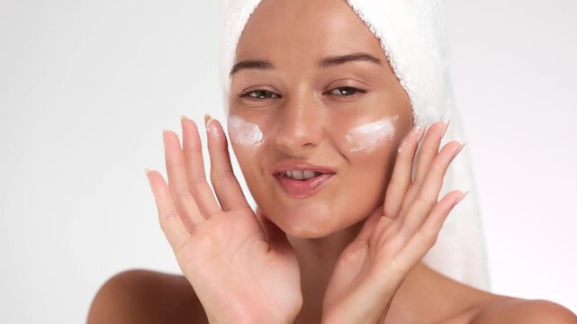 A smiling woman with a towel wrapped around her head playfully makes a kissy face while applying skincare cream to her face. Perfect for illustrating themes of beauty, self-care, and the joy of