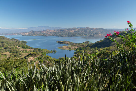 Scenic view of Lago de Suchitl&aacute;n surrounded by lush green hills, vibrant bougainvillea, snake plants and a serene blue sky in Suchitoto, El Salvador. Perfect for lake, nature and travel themes.