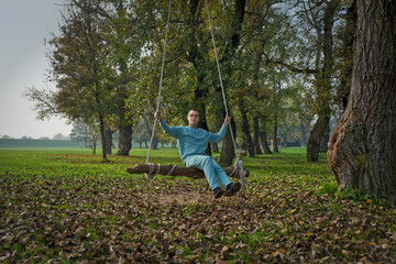 A men enjoys a tranquil moment sitting on a wooden swing suspended by ropes from a tree in a park. The ground is blanketed with fallen leaves