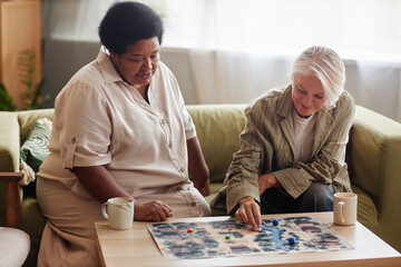 Front view portrait of two senior women playing board game together on coffee table in cozy living...