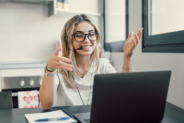 Happy millennial female in headphones watch webinar on laptop in modern office, smiling young businesswoman have fun laugh talk on video call with client or colleague use wireless Internet connection.