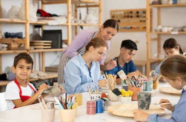 Teenagers and children at a ceramics workshop in an art studio. Woman teacher shows how to paint clay dishes with a brush