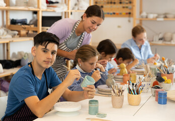 Teenager paints a clay mug with brush at a workshop with a teacher. Boy at a ceramics master class works at a table in an art studio