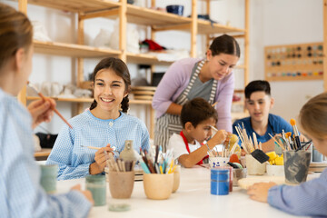 Joyful children, together with the teacher, paint ceramics with brushes at the table. Master class for children on pottery