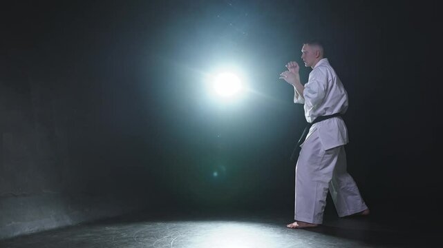 Strong man fighter in white kimono for karate martial arts with black belt preparing for fight training in black background. Man training moves kicks practicing shadow fight ready for sparring