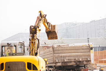 Excavator loading soil into a dump truck on a construction site during the winter season. Snow and...