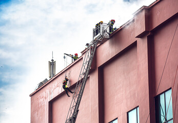 Side view of firefighters extinguish fire on the roof of a high-rise building using extended ladder equipped with water hose to spray water. Firemans extinguishing fire in Nha Trang