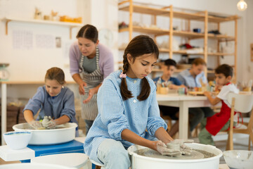Interested preteen girl making clay pot using potter's wheel in art studio together with others