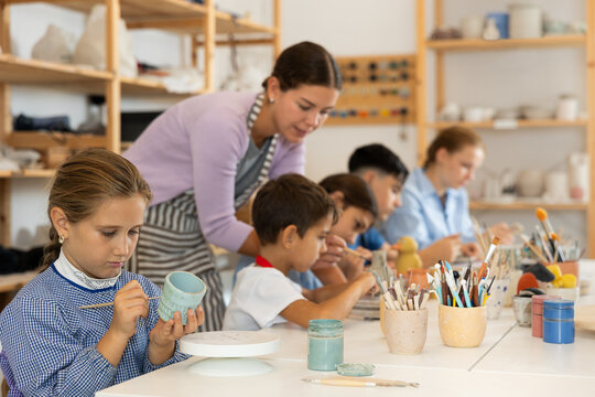 Focused girl paints a ceramic glass made for a master class with a brush. Children make pottery at a workshop in a creative studio
