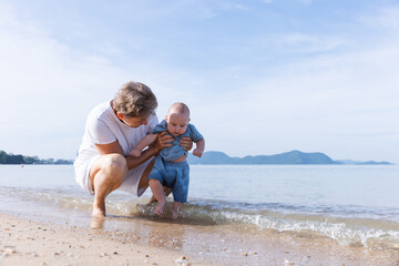 A loving grandmother helps her baby grandchild explore the shoreline, gently dipping the baby's...