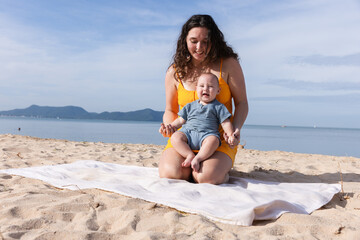 A cheerful mother in an orange dress plays with her laughing baby on a beach towel under the bright...