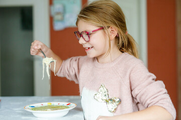 Funny little school girl eat chicken noodles soup with thin pasta. Happy preschool child eating fresh cooked healthy meal with noodles and vegetables at home, indoors.