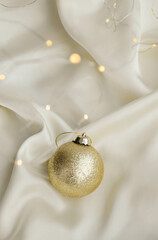 Gold Christmas Ornament on Soft White Fabric. Christmas decoration featuring a glittering golden bauble resting on a smooth white fabric background with soft, warm fairy lights, minimalism and winter-
