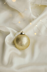 Gold Christmas Ornament on Soft White Fabric. Christmas decoration featuring a glittering golden bauble resting on a smooth white fabric background with soft, warm fairy lights, minimalism and winter-