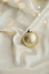 Gold Christmas Ornament on Soft White Fabric. Christmas decoration featuring a glittering golden bauble resting on a smooth white fabric background with soft, warm fairy lights, minimalism and winter-