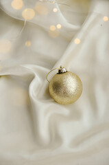 Gold Christmas Ornament on Soft White Fabric. Christmas decoration featuring a glittering golden bauble resting on a smooth white fabric background with soft, warm fairy lights, minimalism and winter-