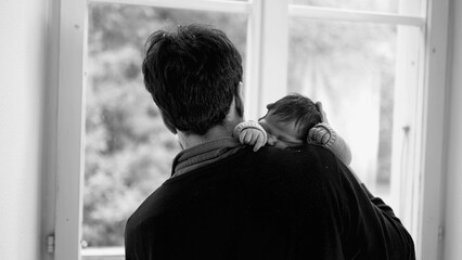 Father gently holding newborn baby over shoulder, gazing out window, bonding moment, family love,...