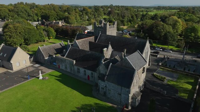 Holy Trinity Abbey Church, Adare, County Limerick, Ireland, September 2024. Drone orbits clockwise in a closeup around the rear of church building as children play in the shade on grassy lawn.