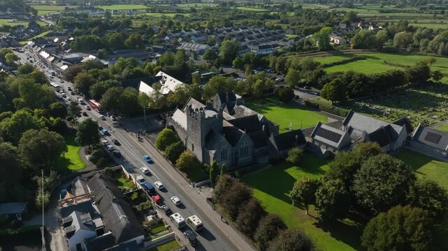 Holy Trinity Abbey Church, Adare, County Limerick, Ireland, September 2024. Drone orbits clockwise above open street to weathered side wall of church building on a bright sunny day.