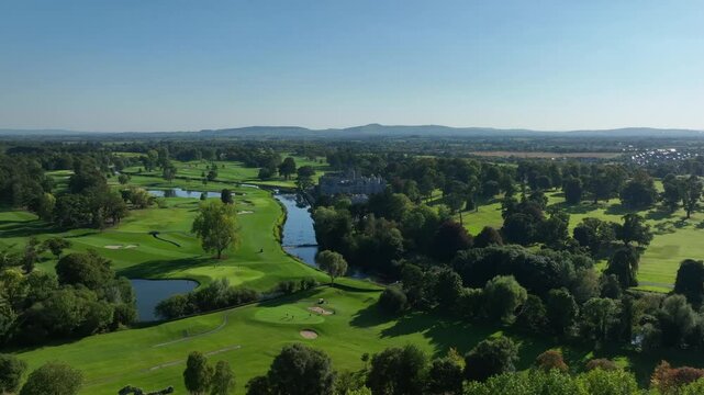 Adare, County Limerick, Ireland, September 2024. Drone rises pulling back while orbiting counter clockwise revealing manor and Franciscan Friary on the river with golf course in a stunning landscape.