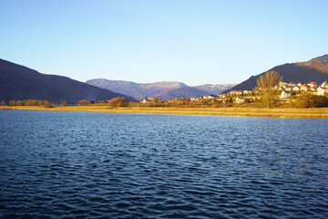 Landscape from the town of Plav: bright blue waters of the lake, thickets of golden reeds, trees with autumn leaves, houses and mountain slopes. Travel to the east of Montenegro: recreation in nature.