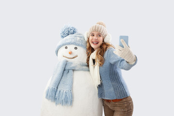 Obraz premium Happy young woman taking a selfie with a snowman, dressed in warm knitted winter clothing. A festive ski trip during the Christmas holidays in the winter season. Social media influencer travel concept