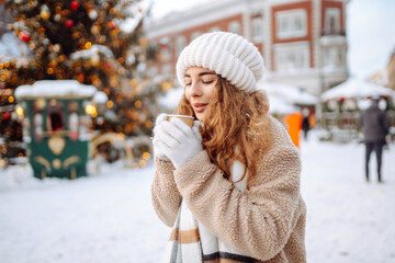 Happy woman drinks a hot drink at a Christmas market decorated with festive lights. Winter holidays, vacation, tourism concept.