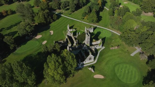 Franciscan Friary, Adare, County Limerick, Ireland, September 2024. Drone orbits clockwise while descending towards the light side of the ruins with walls covered with ivy in middle of a golf course.