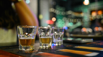 A close-up of three shot glasses with golden liquid on a bar table, set against a vibrant bokeh background of city lights and nightlife ambiance