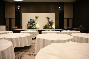 A sophisticated banquet hall with round tables covered in white tablecloths and a stunning floral arrangement backdrop ready for an weddings or formal occasions event