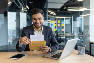 Business professional in office opens envelope, smiling contents. Laptop on desk signify multitasking and technology use. Office setting hints at workspace, possibly involving finance, communication