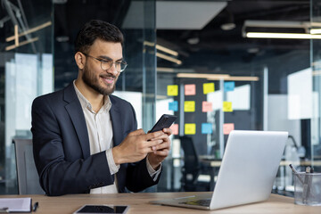Smiling businessman using smartphone in modern office setting with laptop open on desk. Display of productivity, technology integration, and professional communication. Ideal for business themes.