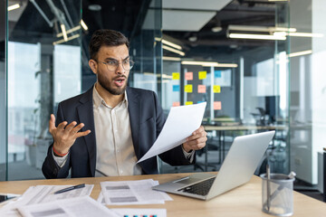 Businessman with shocked expression holding paper, shocked by unexpected report in office. Concept of financial stress, business analysis, and uncertainty. Glass office and laptop visible.