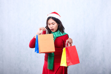 Asian young woman shocked to see the contents of the bag, both hands lifting colorful bags, wearing red christmas sweater with Santa hat and scarf, isolated on white background