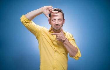 Man shows frame with her fingers. Friendly guy set up definition over blue background, dresses in...