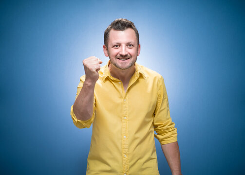 Angry guy looking at camera and shows fist over blue background, dresses in yellow shirt. Frustrated man