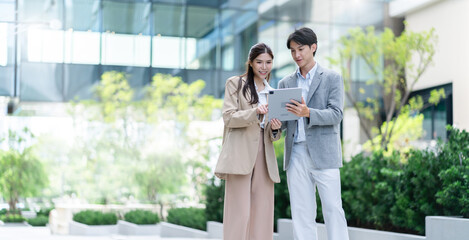 Two asian coworkers standing outside in front of office buildings discuss about business plan and...